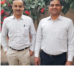 Two men in white shirts standing side by side in front of a flowering plant