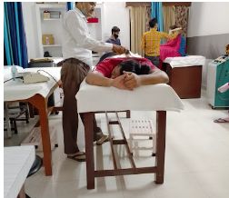 A therapist standing beside a patient lying face down on a wooden treatment couch, with other couches behind