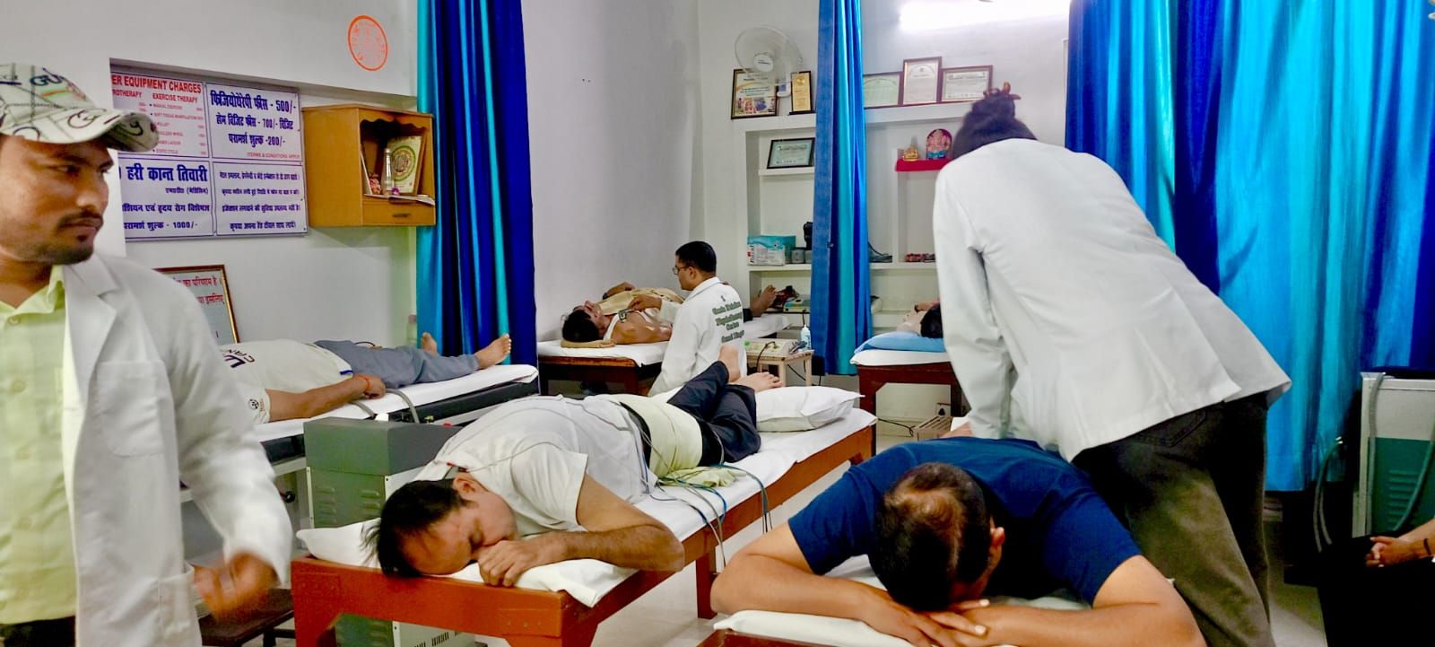 A therapist working on a patient lying face down while other patients rest on couches with electrotherapy leads attached